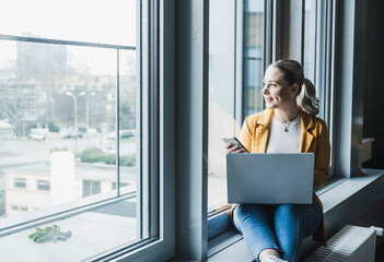 Young businesswoman with smart phone and laptop looking out of window at office
