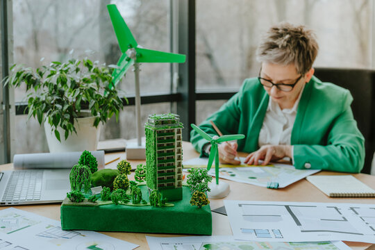 Architect working on blueprint by building and wind turbine models at table in office