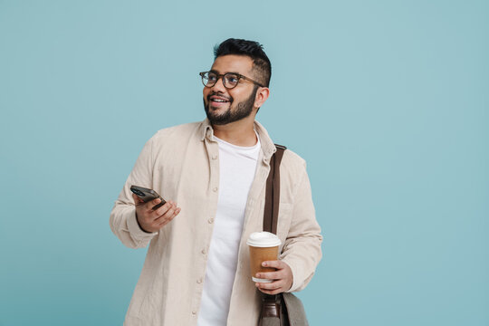 Indian Man Using Mobile Phone And Drinking Coffee While Standing With Bag Isolated Over Blue Wall