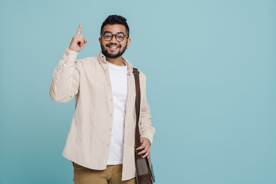 Positive indian guy pointing finger up at copy space while standing with bag isolated