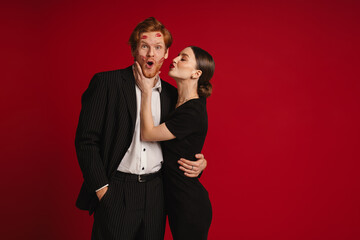 Woman kissing man with red lipstick marks on his face isolated over red wall
