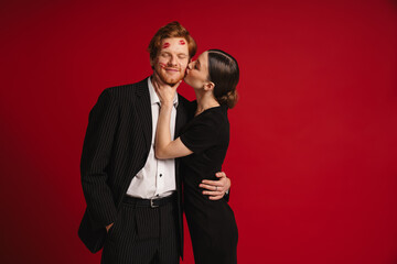 Woman kissing man with red lipstick marks on his face isolated over red wall