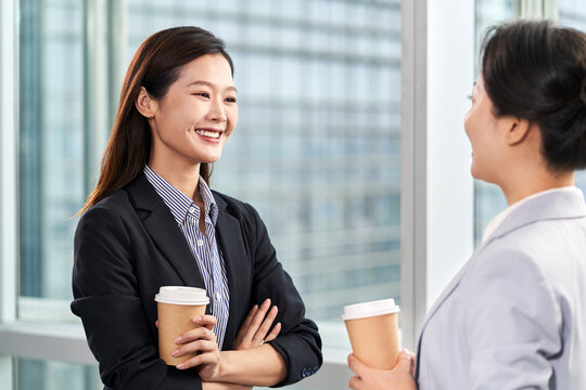 two young asian businesswomen standing chatting talking by the window in office