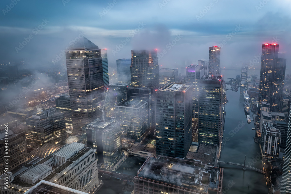 Wall mural The illuminated skyline of Canary Wharf, London, during a foggy evening with the tops of the skyscrapers looking out of the clouds