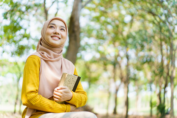 Happy cheerful Asian beautiful female muslim student holding a book and looking and smiling to...