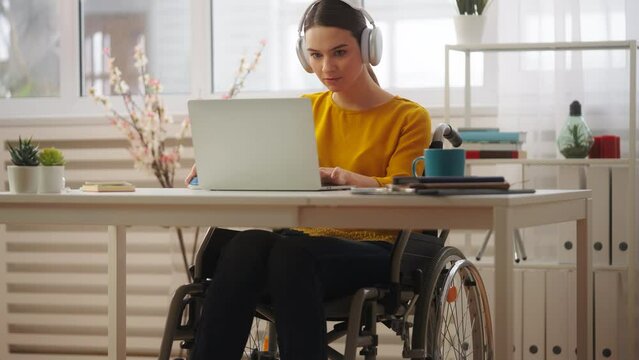 Young woman wheelchair user playing video game on laptop, wearing headset