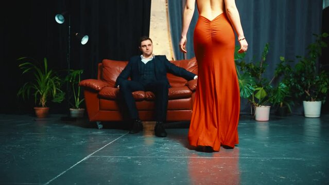 Sexy stylish couple young man in business suit and luxurious woman in evening red dress bare open back. Man sits on leather sofa in dark loft style room. Guy and girl in love together fashion model 4k