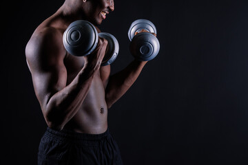 Confident young man shirtless portrait training with dumb-bells against black background.