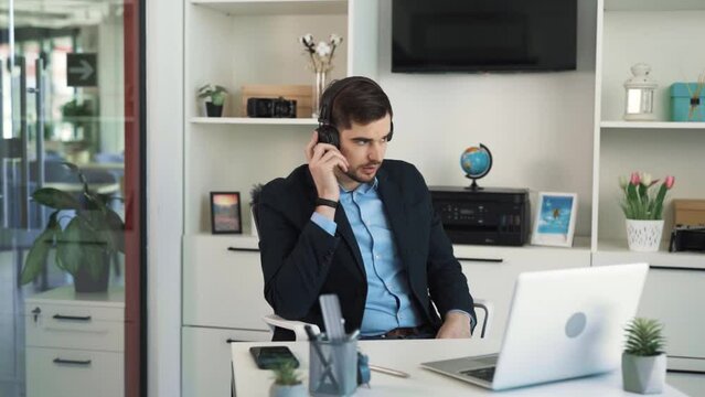 The young attractive man is listening to a new song in his headphones in a bright office. After hearing part of the melody, he begins to adore the song and starts dancing, enjoying the music.