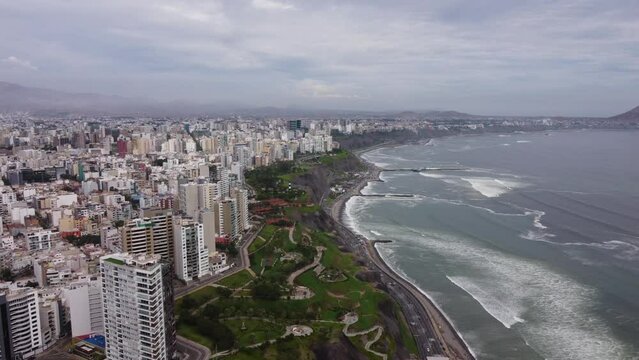 Coastal area called "Costa Verde" with green hills, tall buildings to the left and pacific ocean on the right. Drone flies forward towards the horizon. Located in Miraflores district of Lima, Peru.