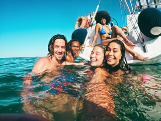 Happy multiracial friends doing selfie swimming in the sea with sail boat in background - Soft focus on center girl face - Travel concept