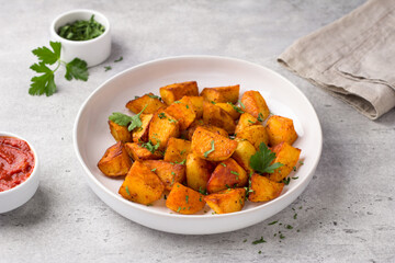 Traditional Spanish potato, patatas bravas with smoked paprika, spicy tomato sauce and parsley in a white bowl on gray stone background, top view. Delicious homemade food