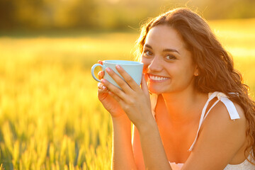 Woman in a field looking at you drinking coffee