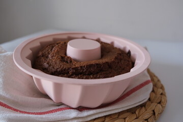 Baked cake in a pink mold, there is a baked cake in a pink round silicone mold on a white kitchen cloth and a placemat on the kitchen table. The concept photo of homemade food.
