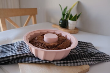 Baked cake in a silicone mold, baked cake with cacao in a pink round silicone mold on dark colored checked kitchen cloth on a white kitchen table. The concept photo of homemade food. 
