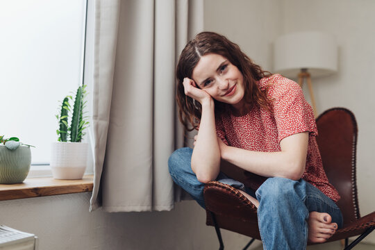 A young adult woman sitting indoors, smiling with an emotion of contentment. She has brown hair in a casual hairstyle and is enjoying her domestic lifestyle.
