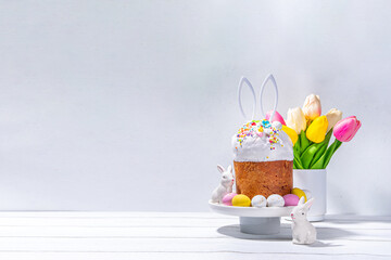 Tasty Glazed Easter cake with sugar decor, colorful eggs basket, ceramic rabbits and spring flower on white kitchen table, copy space. Happy Easter Holidays greeting card background copy space