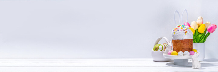 Tasty Glazed Easter cake with sugar decor, colorful eggs basket, ceramic rabbits and spring flower on white kitchen table, copy space. Happy Easter Holidays greeting card background copy space