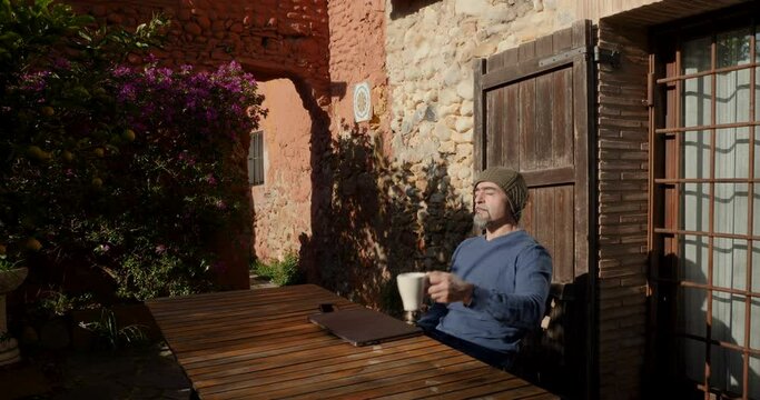 A man close his laptop outside a cottage and drinks coffee enjoying the sun