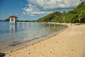 Dreamlike idyllic beach of Coron in the Philippines with palm trees along the beach, a pier leading into the water and hills in the background.