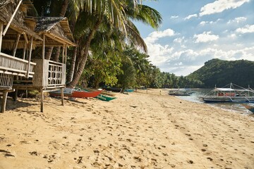 Dreamlike idyllic beach of Coron in the Philippines with palm trees and wooden huts along the beach and hills in the background.