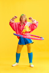 Elderly, cheerful, sportive woman in colorful uniform training, twisting hoop against yellow studio background. Concept of sportive lifestyle, retirement, health care, wellness. Ad