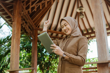 female teacher in hijab with fists clenched looking at the screen of a tablet against a gazebo in...