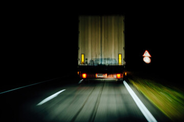 Driving in the darkness, a cargo truck is illuminated by its bright lights as it travels to its destination, leaving behind a trail of reflections and light.