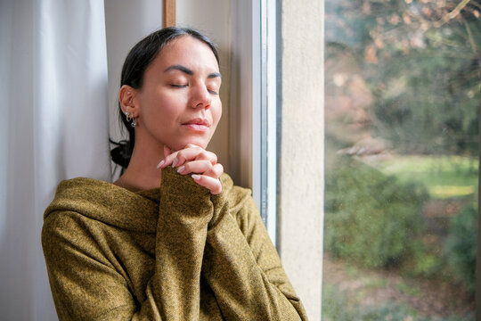 Portrait of a Latina woman in a moment of prayer at her home. She is standing or sitting in front of a window.