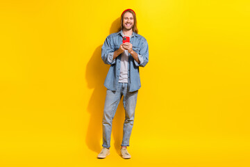 Fototapeta na wymiar Full size photo of good mood man wear denim jacket jeans trousers shoes hold smartphone writing post isolated on yellow color background