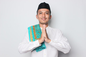 Smiling young Asian man wearing muslim shirt gesturing traditional greeting isolated on white...
