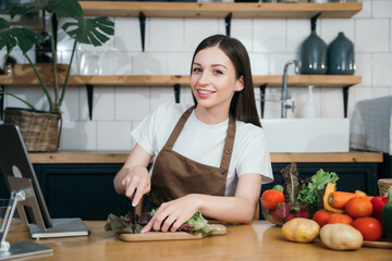 Young beautiful woman using laptop computer searching and learning for cooking healthy food from fresh vegetables and fruits in kitchen room.