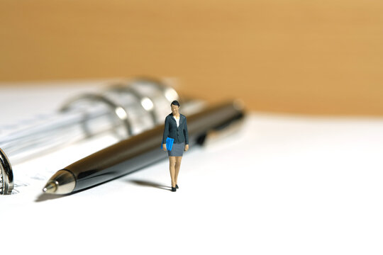 Miniature People Toy Figure Photography. Minutes Of Meeting, Schedule, Task Checklist Concept. A Businesswoman Standing Walking Above Notebook With Pen. Isolated On White Background