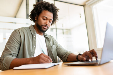 Serious concentrated young african-american businessman, ceo, manager working on laptop, doing online market research, working on project, creating plan or checking financial statement, taking notes