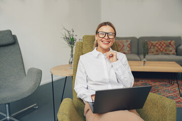 Smiling woman sales manager sitting in cozy chair and working on laptop on coworking background