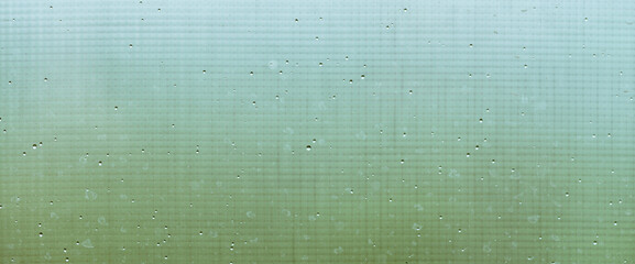 Mosquito net on wet window with rainy drops and dirt spots closeup. Rain droplets on glass with mosquito screen. Blurry minimal background with greenery bokeh against dirty window glass with raindrops