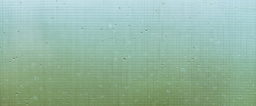 Mosquito net on wet window with rainy drops and dirt spots closeup. Rain droplets on glass with mosquito screen. Blurry minimal background with greenery bokeh against dirty window glass with raindrops