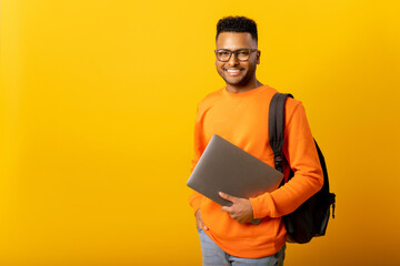 Happy excited foreign male student with backpack standing isolated on yellow and holding laptop,...