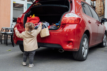 Family road trip where toddler little girl is helping to load up the trunk with suitcases and backpacks. Little girl help and put bags into the car trunk