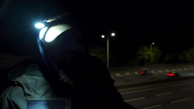 A dark view of a bearded man talking beside a motorway wearing a hood and a headtorch