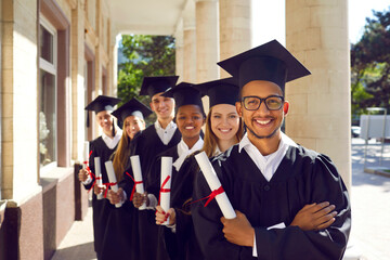 Groups of happy and joyful graduates in academic gowns and academic caps holding scrolls of...