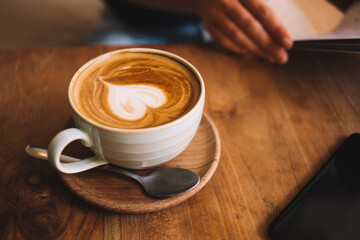 Cropped image of ceramic cup and spoon with aroma caffeine beverage for energy and awaken on wooden table, top view image of late art on milky foam on coffee beverage for breakfast