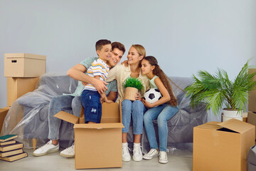 Fototapeta na wymiar Happy friendly family with children are moving in new modern apartment house. Mom, dad and daughter are sitting on couch and son is standing in box near. They are smiling and cuddling each other.