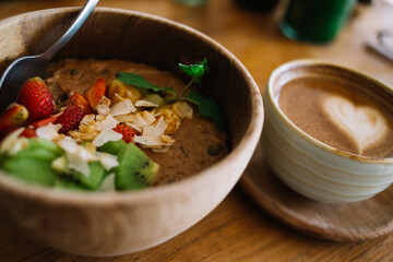 Soft focus. Close up picture of wooden buddha bowl with ripe vitamin fresh fruits for healthy low fat high fiber nutrition, cropped image of coffee cup with milky foam and tasty food for breakfast