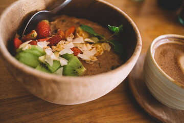 From above of delicious healthy smoothie bowl with berries kiwi and coconut placed on wooden table near cup of hot coffee