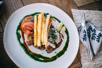 Top view of meat dish with vegetables and French fries seasoned with green sauce and fork on napkin on table