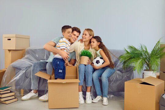 Happy friendly family with children are moving in new modern apartment house. Mom, dad and daughter are sitting on couch and son is standing in box near. They are smiling and cuddling each other.