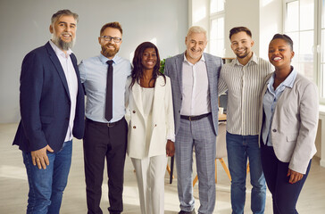 Fototapeta premium Group of business multiethnic people men and women looking at camera hugging and smiling indoors. Company employees team or group of staff standing in the office confidently and cheerfully.