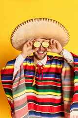 Fresh. Young funny man in colorful festive clothes, poncho and sombrero posing with lime near eyes against yellow studio background. Concept of mexican traditions, fun, celebration, festival, emotions