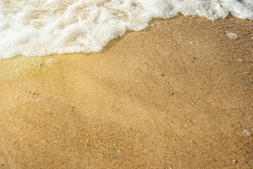 Close-up of the sea wave with white foam flowing onto the sandy beach. Background with crystal clear water on the theme of seaside holidays and family weekends on the beach.